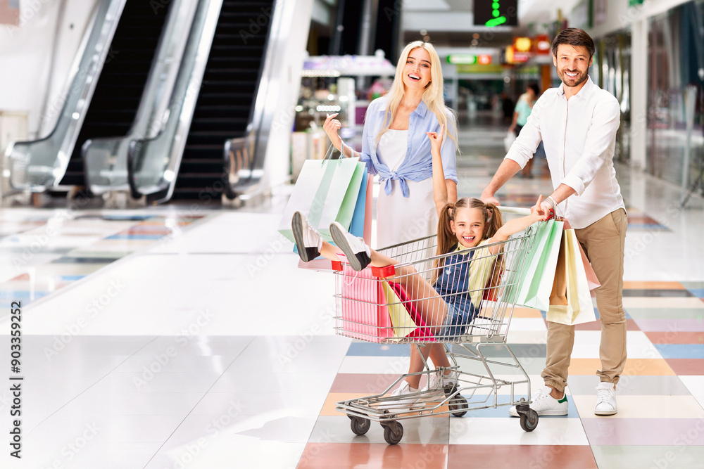 Obraz premium A young girl sits in a shopping cart being pushed by her father, while her mother walks behind with shopping bags. The family is smiling and laughing as they navigate the modern mall