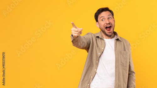 Photos A man in a brown shirt stands in front of a yellow background, pointing his index finger at copy space and looking excitedly in that direction, with his mouth open in a smile