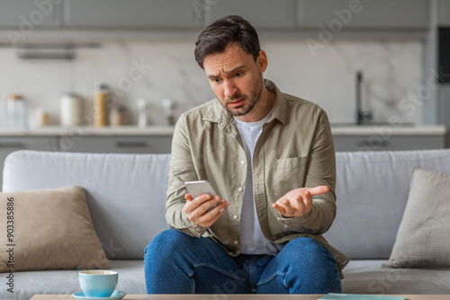 A man sits on a couch in his living room, holding his phone with a confused expression on his face. He gestures with his other hand, as if trying to explain or clarify something.