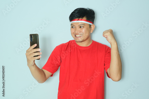 Indonesian man clenched fist showing excitement when looking to his smartphone. Wearing a red t-shirt