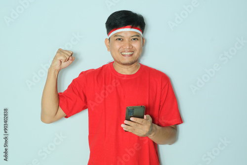 Indonesian man smiling while holding a smartphone during independence day celebration. Wearing a red t-shirt
