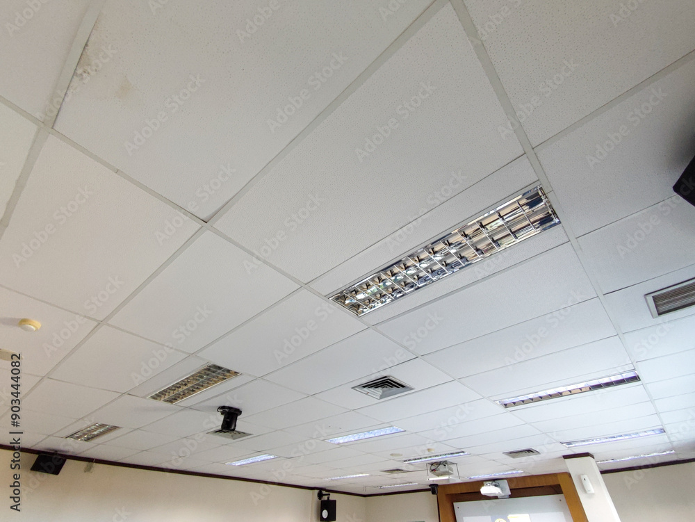 Office ceiling with white acoustic panels, fluorescent lights, air vents, and signs of water stains. Aged infrastructure shows discoloration, dust accumulation, and minor damage on the suspended tiles