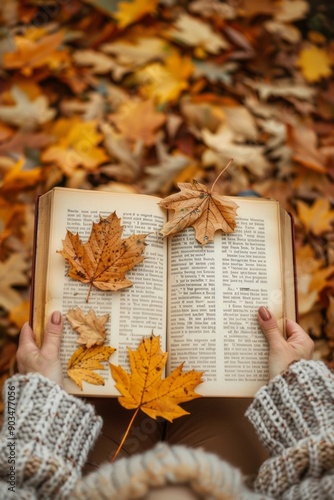 An open book with yellow and brown autumn leaves scattered on the pages, held by a person in a knitted sweater, set on a bed of fallen leaves providing a nostalgic autumn feel. photo