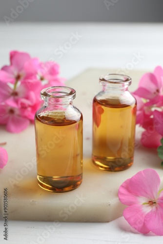 Bottles of geranium essential oil and beautiful flowers on white table, closeup