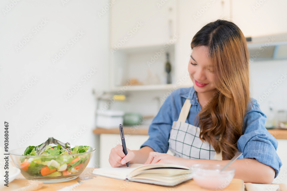 Young asian woman taking notes about food recipes in notebook and eating yogurt while preparing vegetables salad and cooking healthy breakfast food in modern kitchen with healthy lifestyle at home Young asian woman taking notes about food recipes in notebook and eating yogurt while preparing vegetables salad and cooking healthy breakfast food in modern kitchen with healthy lifestyle at home