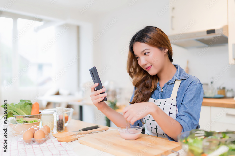 Young asian woman eating yogurt and use smartphone to searching food recipes while preparing fresh vegetables salad and cooking healthy breakfast food in modern kitchen with healthy lifestyle at home Young asian woman eating yogurt and use smartphone to searching food recipes while preparing fresh vegetables salad and cooking healthy breakfast food in modern kitchen with healthy lifestyle at home