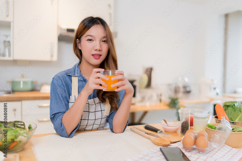 Young asian woman holding glass of orange juice and drinking fresh fruits while preparing vegetables salad and cooking healthy breakfast food in modern kitchen with healthy lifestyle at home Young asian woman holding glass of orange juice and drinking fresh fruits while preparing vegetables salad and cooking healthy breakfast food in modern kitchen with healthy lifestyle at home
