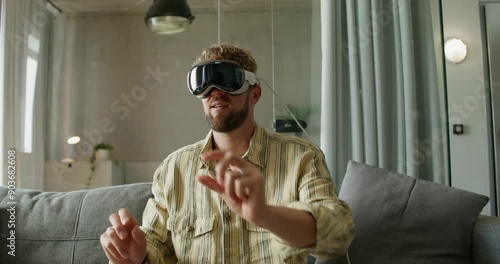 A man in a virtual reality headset moves his hands while sitting on a sofa in a home interior, daylight