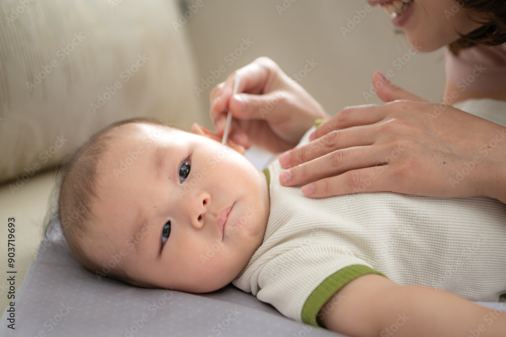 A tiny child smiles happily on the bed and mother is using a cotton bud to clean child ears A tiny child smiles happily on the bed and mother is using a cotton bud to clean child ears