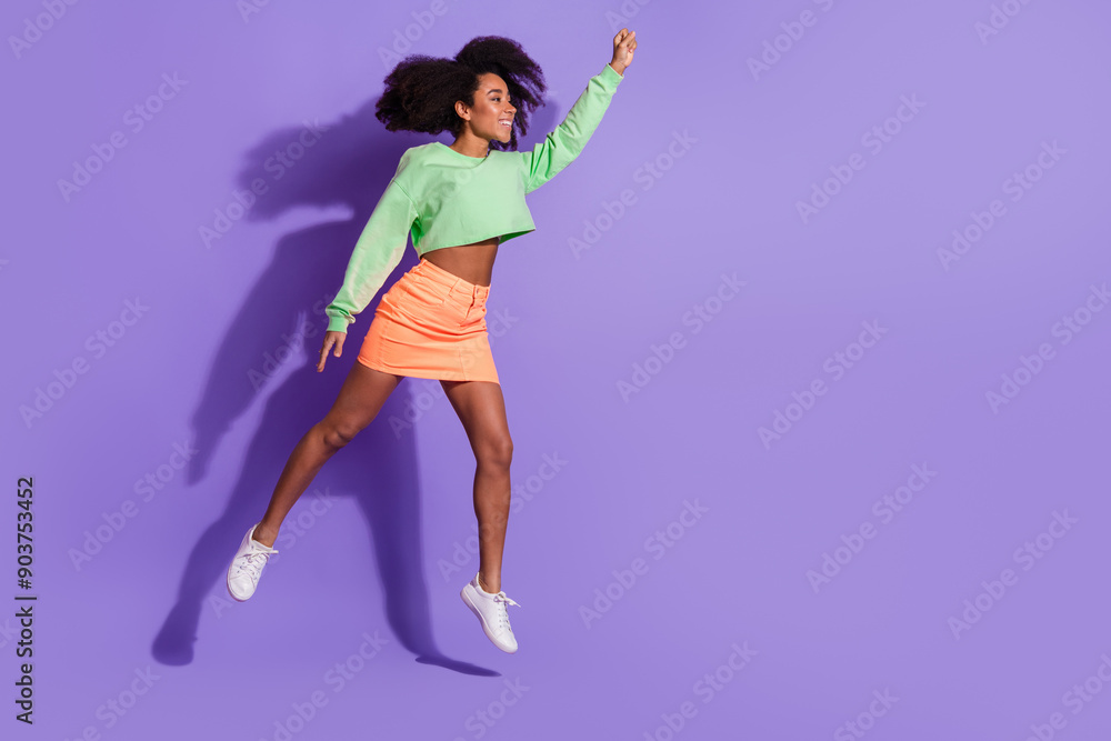 Full body length photo of young funny jumping girl with wavy hair in green top raised fist up superhero isolated on violet color background Full body length photo of young funny jumping girl with wavy hair in green top raised fist up superhero isolated on violet color background