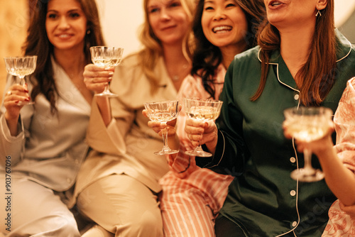 Happy girls with glasses of champagne laugh and raise a festive toast. Young smiling women of different races in pajamas drink sparkling wine at a house party. photo