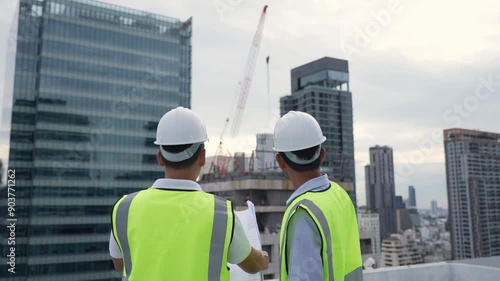 Two engineer men in safety vests are standing on a rooftop of a building. They are looking at something and discussing it