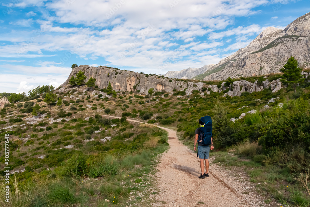 Obraz premium Hiker man with baby carrier backpack on idyllic hiking path to Tucepi town along Makarska Riviera, Dalmatia, Croatia, Europe. Majestic Biokovo mountains rise up against dramatic sky. Dinaric Alps
