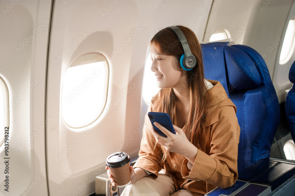 An Asian woman travels abroad by plane, sitting near the window. She listens to music while flying, looks at her phone, uses a tablet, and smiles happily, enjoying her trip. An Asian woman travels abroad by plane, sitting near the window. She listens to music while flying, looks at her phone, uses a tablet, and smiles happily, enjoying her trip.