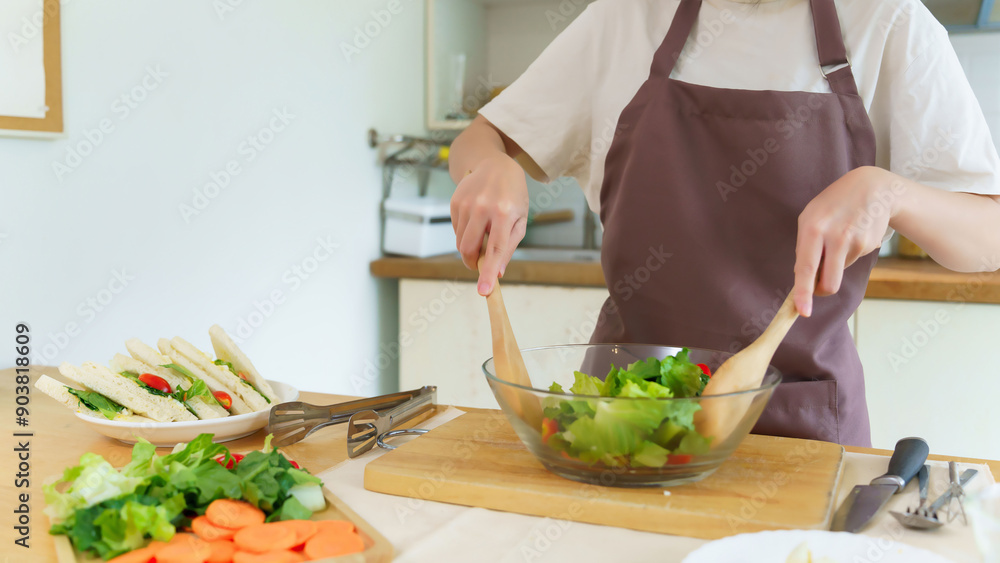 Asian women using wooden spoon to mixing ingredients and slices of fresh vegetable in the glass bowl to cooking breakfast meal and vegetable salad while making homemade healthy food lifestyle in home