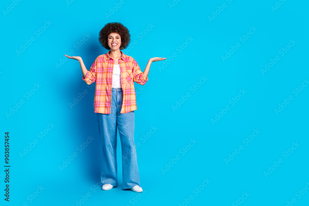 Full length photo of lovely young lady compare hold empty space dressed stylish checkered garment isolated on blue color background Full length photo of lovely young lady compare hold empty space dressed stylish checkered garment isolated on blue color background