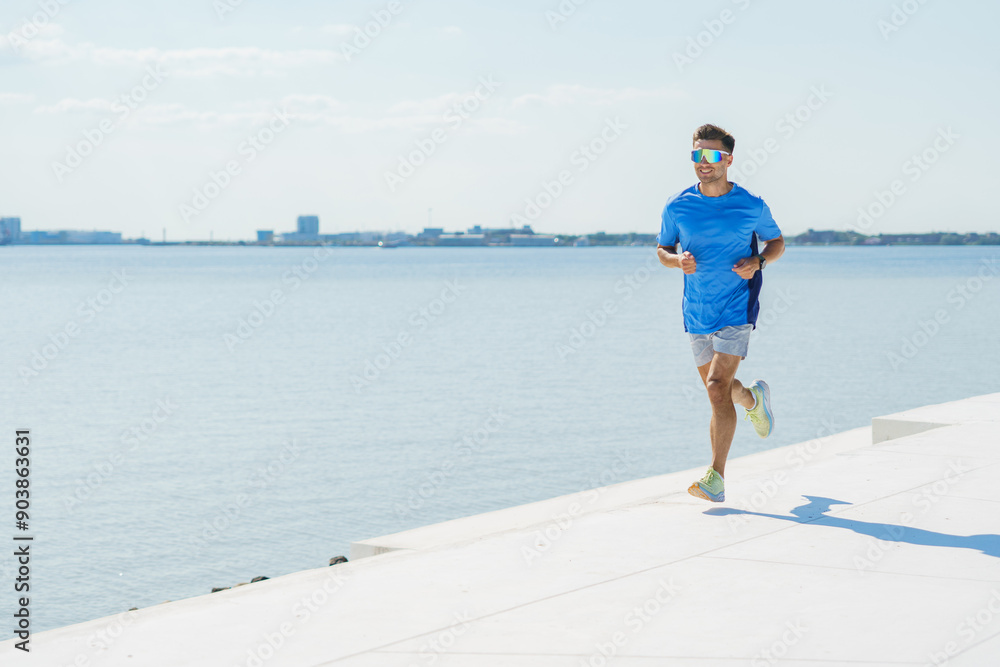 A man in a blue shirt runs along a waterfront, enjoying the clear day and scenic views. A man in a blue shirt runs along a waterfront, enjoying the clear day and scenic views.