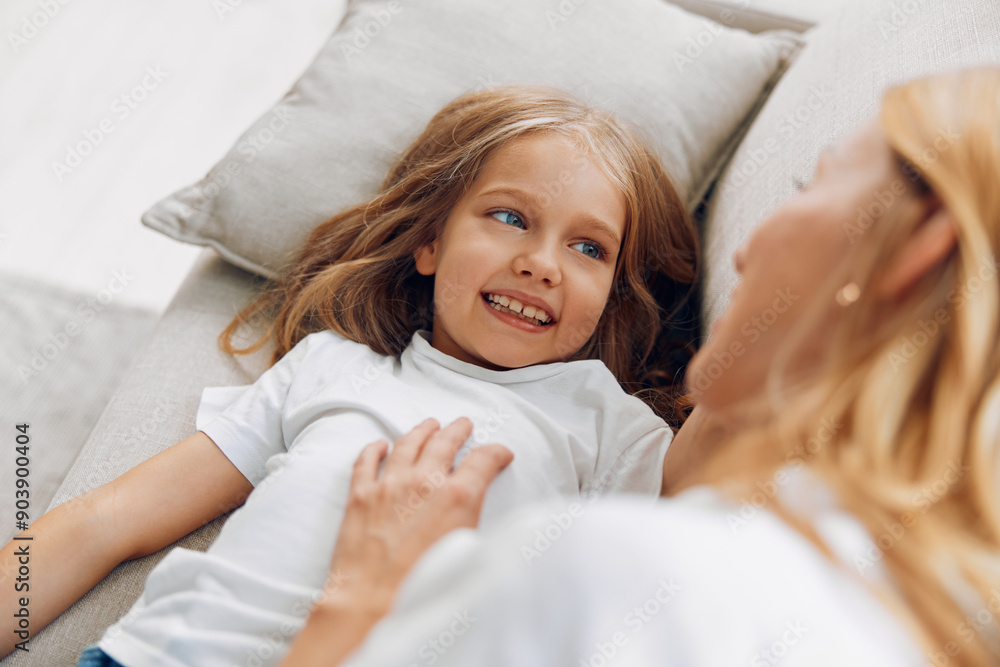 Smiling little girl relaxing on couch with her mother, sharing a happy moment in a cozy home environment Smiling little girl relaxing on couch with her mother, sharing a happy moment in a cozy home environment