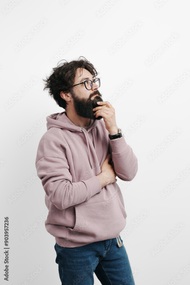 A pensive man with a beard and glasses wearing a casual hoodie stands with his hand on his chin, looking contemplative against a white background. A pensive man with a beard and glasses wearing a casual hoodie stands with his hand on his chin, looking contemplative against a white background.