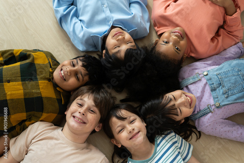 Diverse group of happy children lying on the floor in a circle, smiling and bonding together, showcasing multicultural friendship and unity in a cheerful and playful setting.