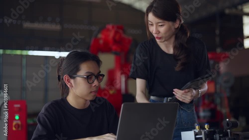 Two engineers collaborating on a project using a laptop and tablet in a factory, with industrial machinery in the background. photo