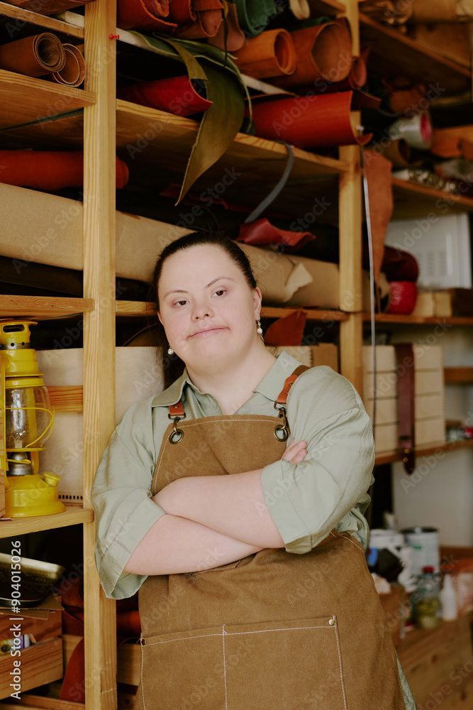 Talented female tailor with down syndrome dressed in t-shirt and chamois apron leaning on wooden shelves in atelier Talented female tailor with down syndrome dressed in t-shirt and chamois apron leaning on wooden shelves in atelier