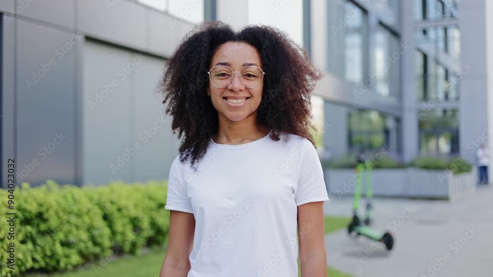 Portrait of cheerful african american woman with clear glasses gazing directly at camera with contemporary building and electric scooter in background. Attractive brunette woman smiling warmly. Portrait of cheerful african american woman with clear glasses gazing directly at camera with contemporary building and electric scooter in background. Attractive brunette woman smiling warmly.