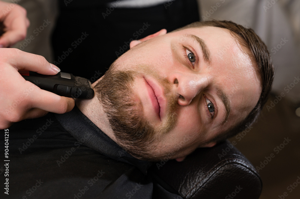 Obraz premium A barber in the salon trims the beard of a man with a electric shaver in the neck area