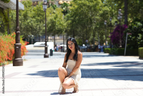 Young Spanish woman, brunette and beautiful in white dress crouching on her knees and doing different postures on a pedestrian avenue in the centre of Seville, Spain. Beauty and fashion concept.