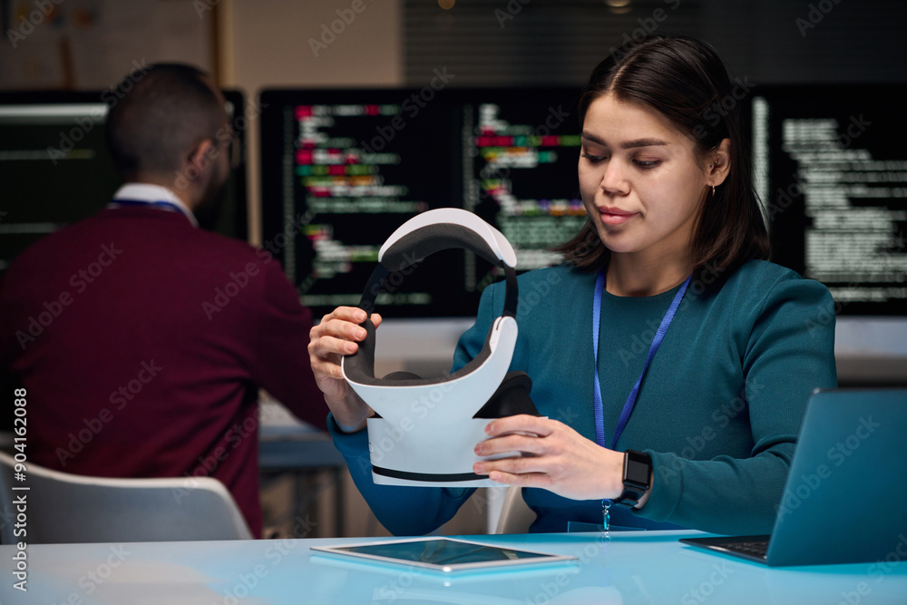 Portrait of young woman setting up VR headset while developing new technology in office with code screens in background, copy space Portrait of young woman setting up VR headset while developing new technology in office with code screens in background, copy space