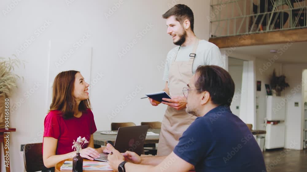 In a cozy and bustling cafe, a barista guy is taking an order from a young couple at the counter. The warm and inviting atmosphere of the cafe creates a pleasant setting as he attentively listens and