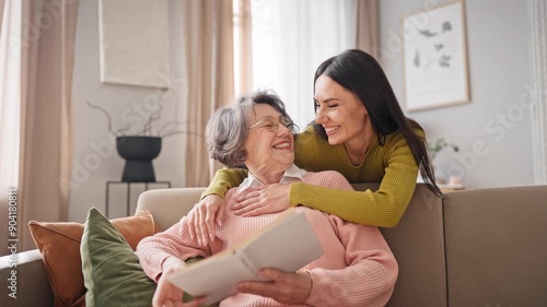 Elderly Caucasian woman with grey hair wearing glasses while reading book. Charming daughter walking up behind mature mother and hugging her. Older mom widely smiling while looking at her relative.