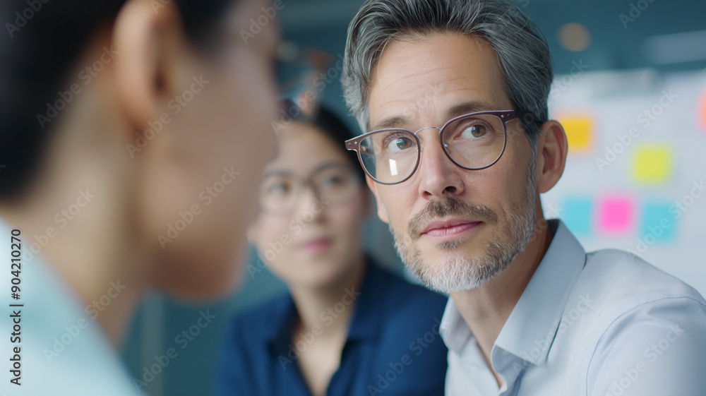 A middle-aged man with glasses engages in discussion with colleagues in a modern office. Blurred sticky notes in the background emphasize brainstorming and collaboration. A middle-aged man with glasses engages in discussion with colleagues in a modern office. Blurred sticky notes in the background emphasize brainstorming and collaboration.