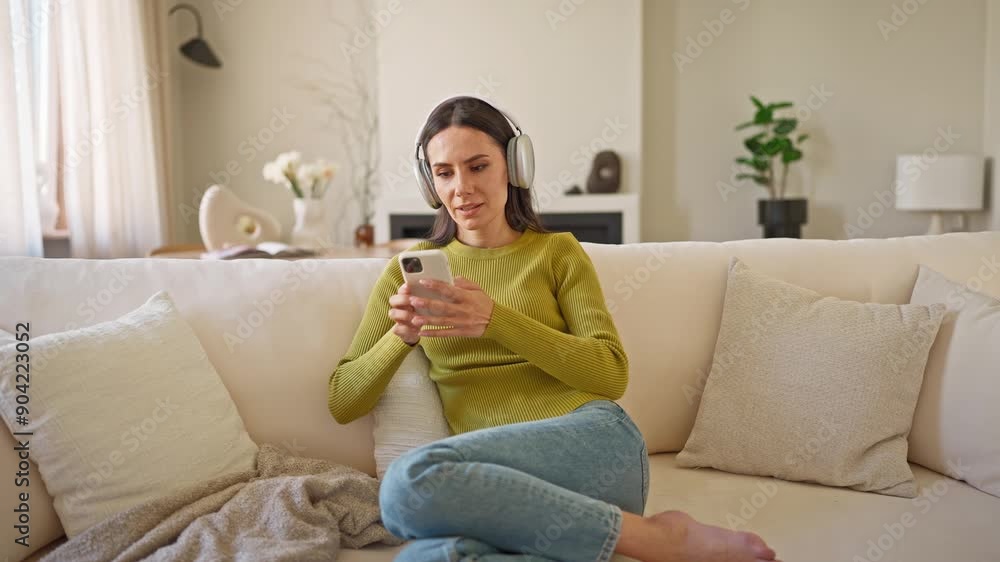 Pretty Caucasian woman relaxing at home. Messaging with friends in app or social media. Using cell phone for wireless communication. Recreation concept. Listening to music while wearing headphones. Pretty Caucasian woman relaxing at home. Messaging with friends in app or social media. Using cell phone for wireless communication. Recreation concept. Listening to music while wearing headphones.