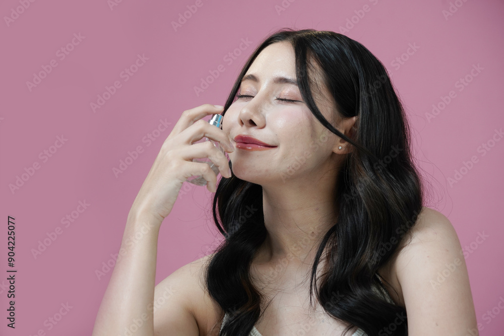 Obraz premium Beautiful Asian woman with Korean style makeup holding a bottle of perfume smelling the fragrance on a pink background