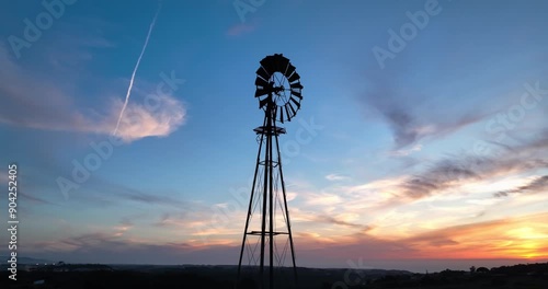 A vintage metal windmill in rural Portugal powers a farm water pump in the dry outback. A drone shot captures blades rotating against a sunset backdrop, showcasing the timeless beauty of this scene.
