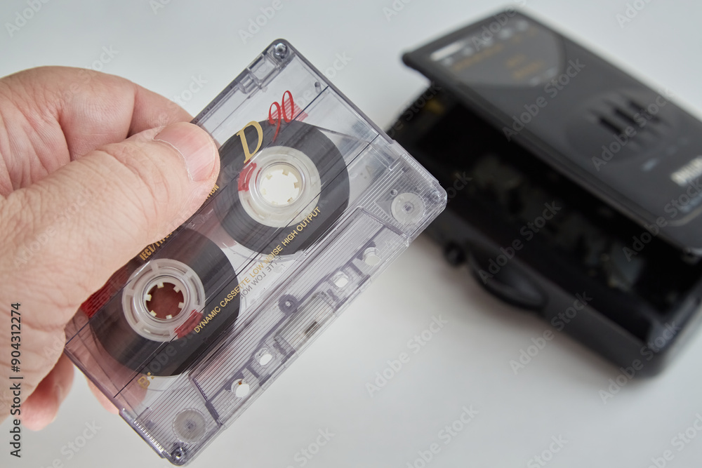 Obraz premium A hand holding an old audio cassette against a white background, with an old audio player in the background, close-up, music from past years, old audio recordings, old analog technologies