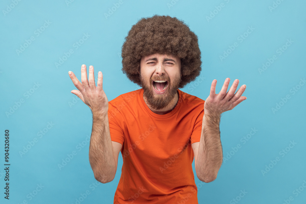 Portrait of man with Afro hairstyle wearing orange T-shirt spreading hands, seriously looking at camera, family quarrel, misunderstanding. Indoor studio shot isolated on blue background. Portrait of man with Afro hairstyle wearing orange T-shirt spreading hands, seriously looking at camera, family quarrel, misunderstanding. Indoor studio shot isolated on blue background.