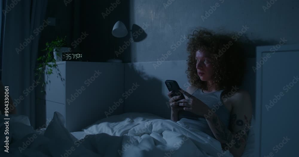 A woman uses a mobile phone sitting in bed. The offended man lies down next to her and turns away. A family quarrel A woman uses a mobile phone sitting in bed. The offended man lies down next to her and turns away. A family quarrel