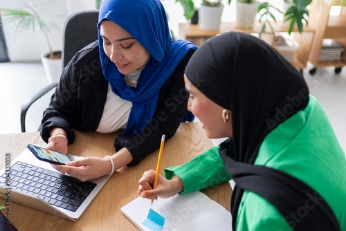 Two women in hijabs effectively planning their goals in a modern office environment, using laptops, notes, and a goal-setting approach. photo