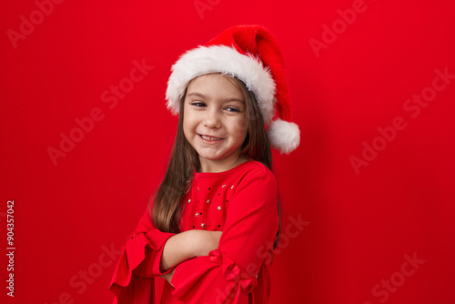 Adorable hispanic girl, arms crossed, dons a christmas hat over red background. her happy, radiant smile beams at the camera. what a bundle of positivity!