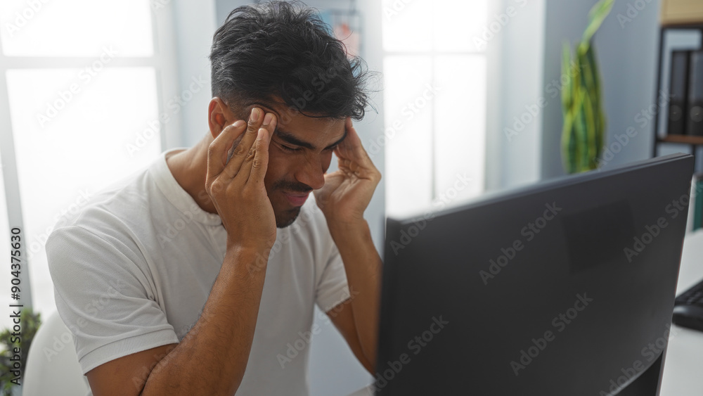 Young hispanic man in an office holding his head in frustration while sitting in front of a computer Young hispanic man in an office holding his head in frustration while sitting in front of a computer