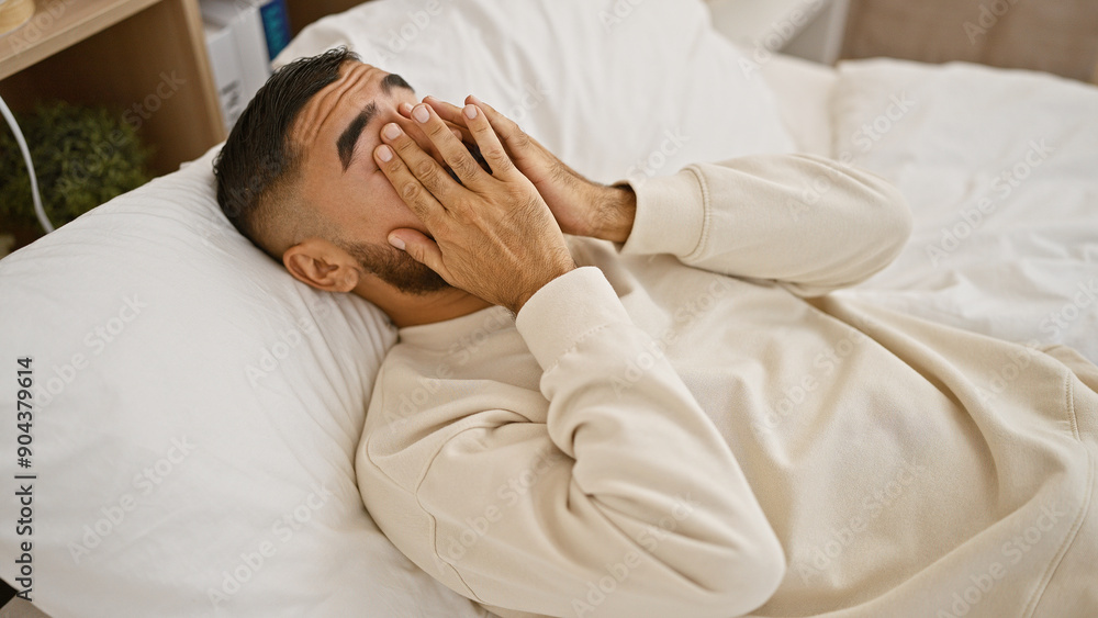 A tired man covers his face with hands lying in bed, suggesting morning frustration or headache in a bedroom setting. A tired man covers his face with hands lying in bed, suggesting morning frustration or headache in a bedroom setting.