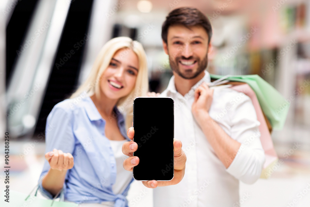 A man and woman, both smiling, stand in a shopping mall with bags, holding up a smartphone with black empty screen A man and woman, both smiling, stand in a shopping mall with bags, holding up a smartphone with black empty screen