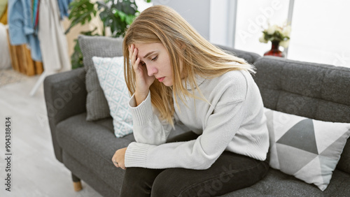 Tableau sur toile A pensive blonde woman with blue eyes wearing a white sweater sits on a gray couch in a modern living room, appearing contemplative or worried