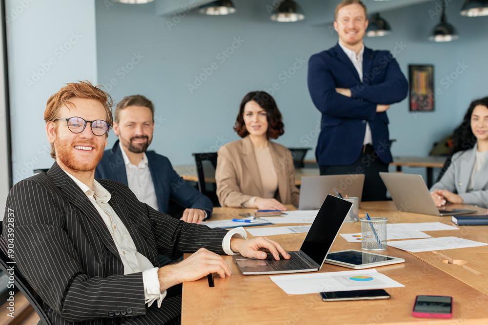 A team of business professionals is gathered around a large conference table, collaborating and working on projects. One man, with a red beard and glasses, is smiling and working on a laptop A team of business professionals is gathered around a large conference table, collaborating and working on projects. One man, with a red beard and glasses, is smiling and working on a laptop