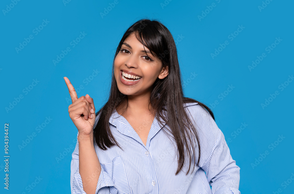 Woman Smiling and Pointing Upward Against a Bright Blue Background in a Casual Outfit Woman Smiling and Pointing Upward Against a Bright Blue Background in a Casual Outfit