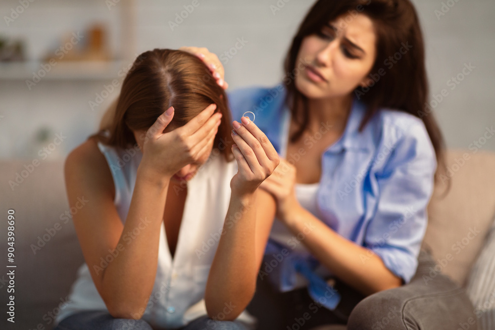 Divorce Concept. Friend Comforting Desperate Girl After Breakup With Ex-Husband Holding Ring Sitting On Couch At Home. Divorce Concept. Friend Comforting Desperate Girl After Breakup With Ex-Husband Holding Ring Sitting On Couch At Home.