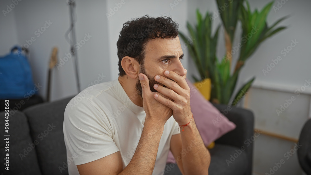 A pensive bearded man sits thoughtfully indoors, hand on face, expressing concern or deep thought in a modern living room. A pensive bearded man sits thoughtfully indoors, hand on face, expressing concern or deep thought in a modern living room.