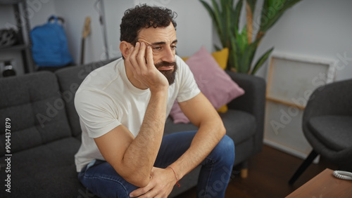 Pensive bearded man in casual attire seated indoors on a cozy sofa in a modernly decorated living room. photo